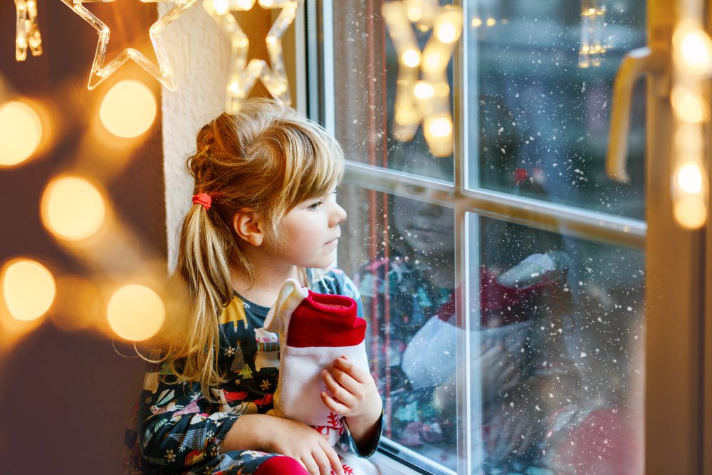 Happy child wait on holiday by window with Christmas lights in winter.